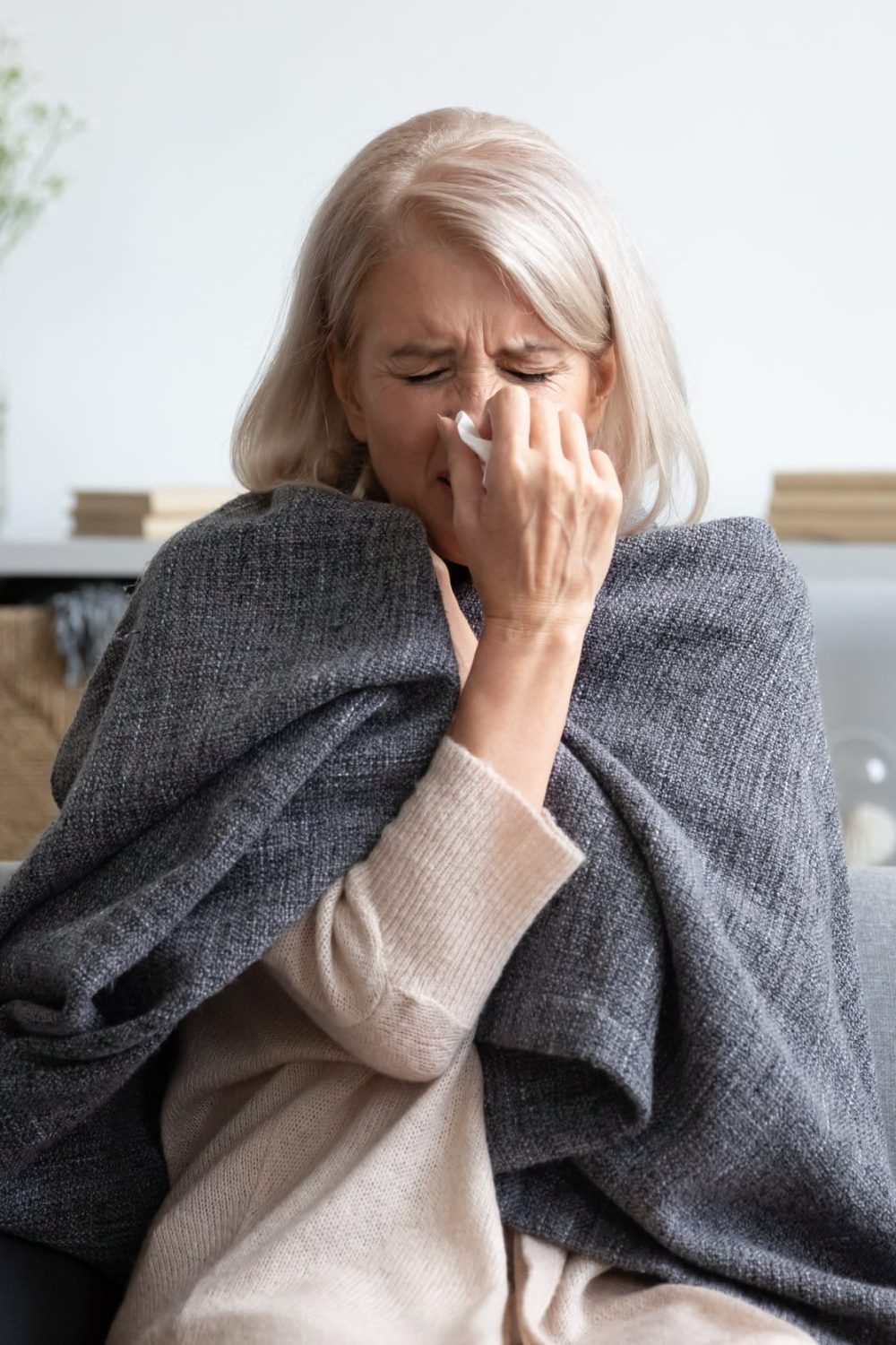 Middle-aged 50s sick frozen woman seated on sofa in living room covered with warm plaid sneezing holding paper napkin blow out runny nose feels unhealthy, seasonal cold, weakened immune system concept